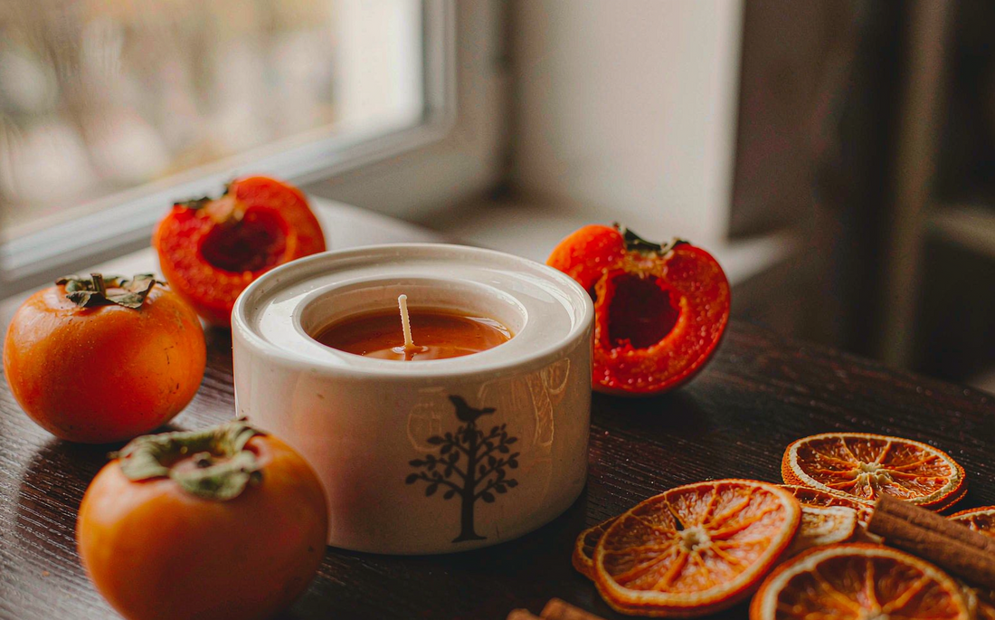 Persimmon candle beside fresh persimmons, dried orange and cinnamon sticks